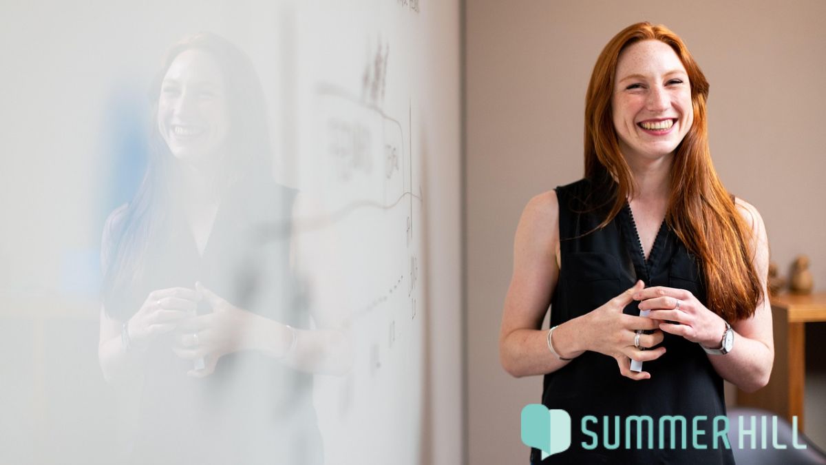 A red-haired woman stands beside a whiteboard, smiling as sunlight falls on her, looking confident and happy while presenting.