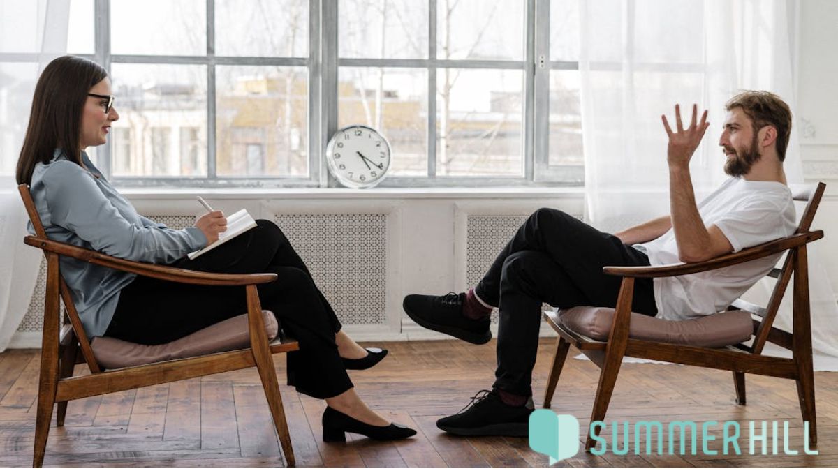 A man speaks with a female therapist in a counseling room, sitting before a large window, both looking animated and focused in discussion.