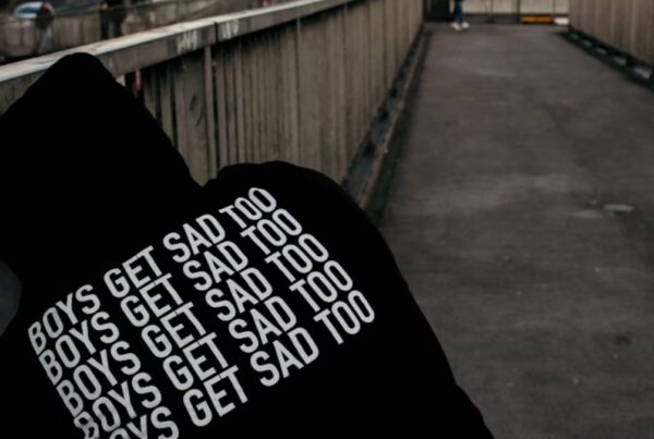 Man wearing a hooded sweatshirt bending down on the street, with the label reading ‘Boys Get Sad Too.’