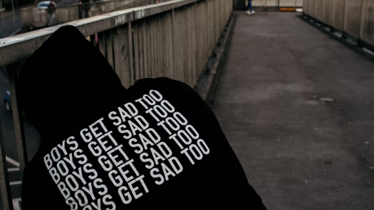Man wearing a hooded sweatshirt bending down on the street, with the label reading ‘Boys Get Sad Too.’