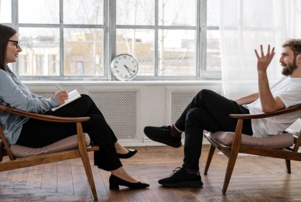 Female therapist listening attentively to a male patient during a discussion, with a large window in front of them.