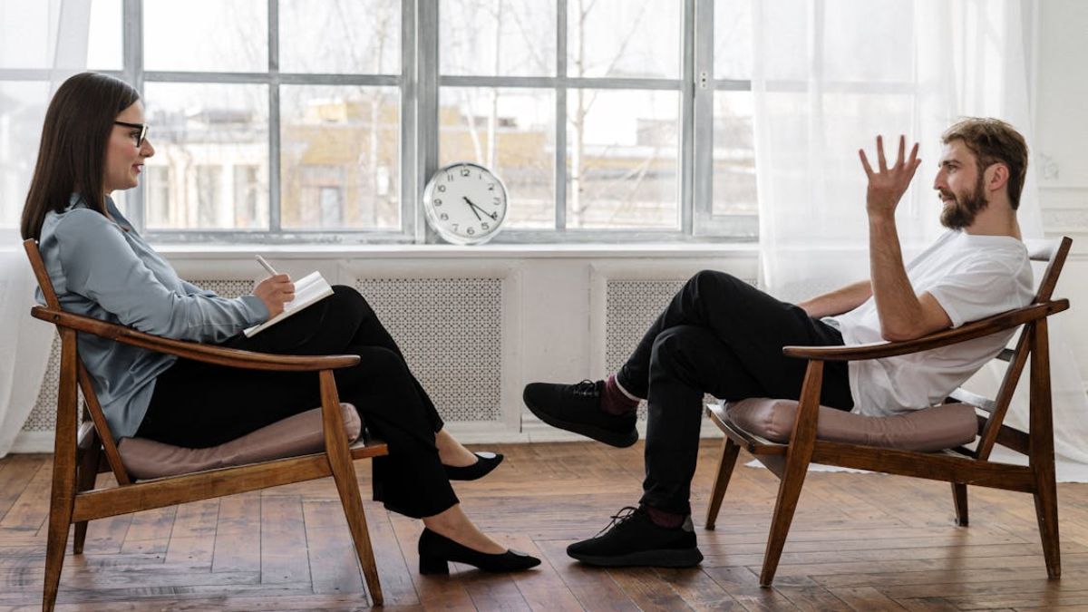 Female therapist listening attentively to a male patient during a discussion, with a large window in front of them.