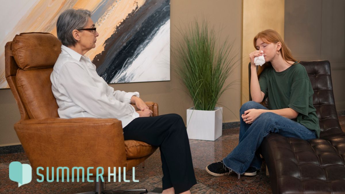 Mental health clinic services, a female patient speaks with her female therapist in a brightly lit room while holding a handkerchief as she becomes emotional.