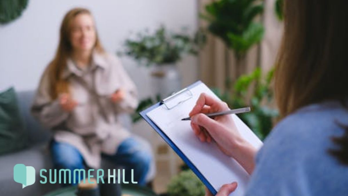 A woman receives CBT for bipolar disorder, shown in soft focus while the therapist takes notes during the session.