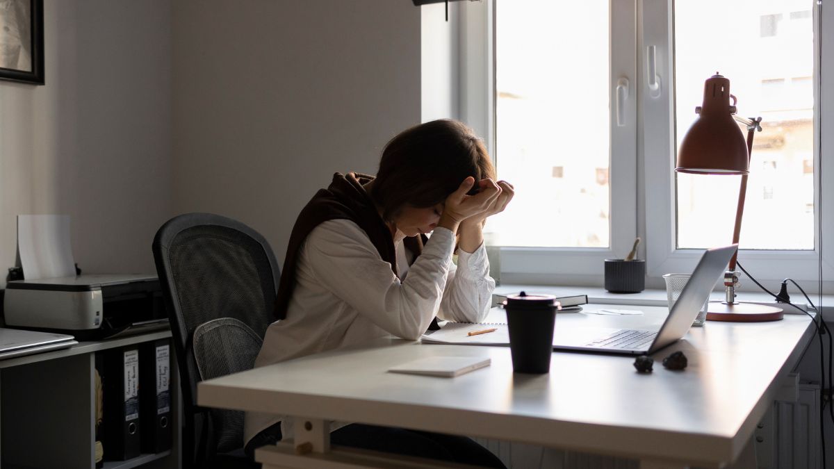Masking Anxiety: A woman sits in an office with a wide window framing the skyline, her hands covering her face in an act of frustration.
