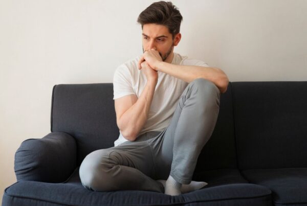 A man sits pensively on a couch in a bright apartment, his elbow resting on his knee and his arms forming a T as he stares ahead, illustrating how therapy can feel hard.