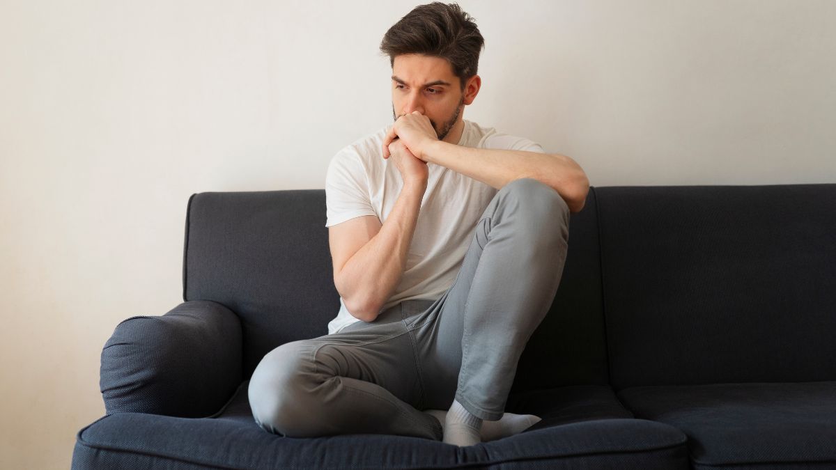 A man sits pensively on a couch in a bright apartment, his elbow resting on his knee and his arms forming a T as he stares ahead, illustrating how therapy can feel hard.