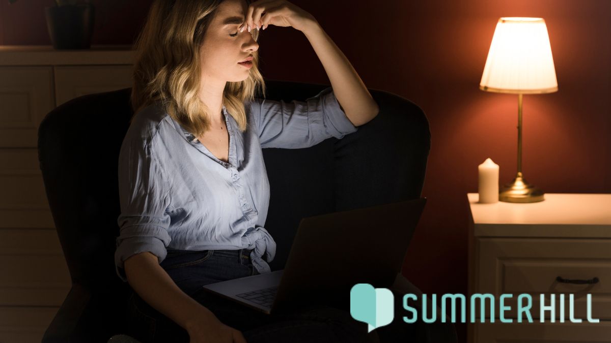 A woman closes her eyes and touches her forehead while sitting in bed, with her bed in the background as she struggles with overthinking at night before bedtime.