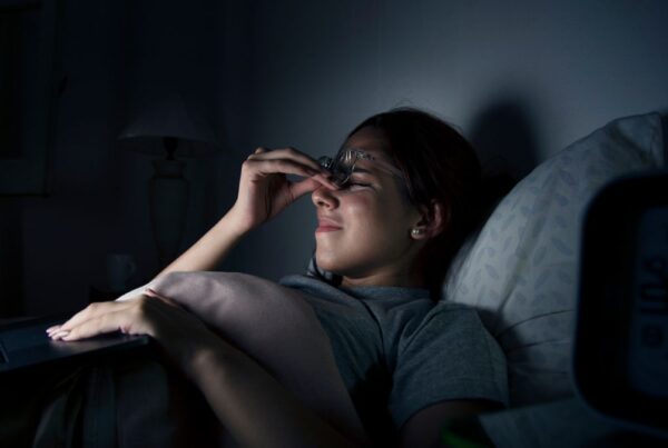 A woman wearing glasses lies in bed beside a night lamp, squeezing her eyes with her hands, unable to sleep from overthinking at night.