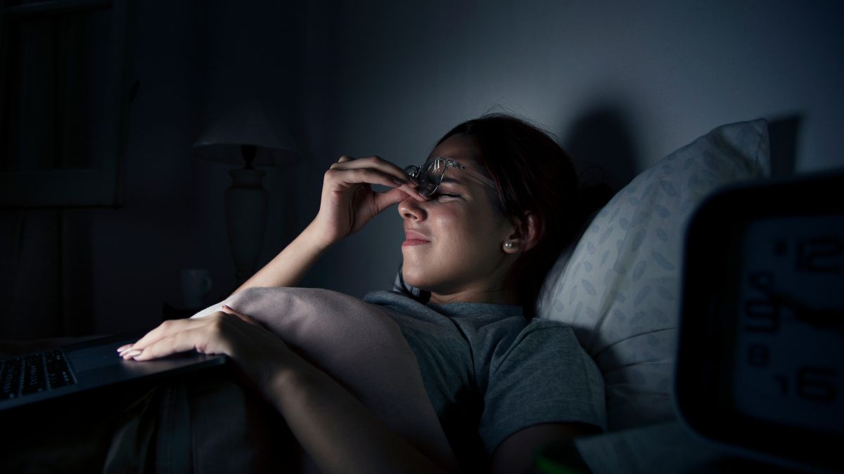 A woman wearing glasses lies in bed beside a night lamp, squeezing her eyes with her hands, unable to sleep from overthinking at night.