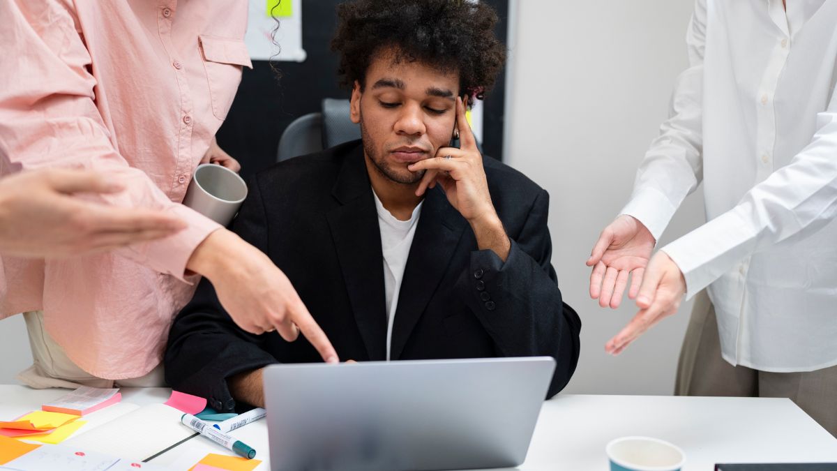 Masking Anxiety: A man sits in an office with a laptop before him, while standing figures point at him.