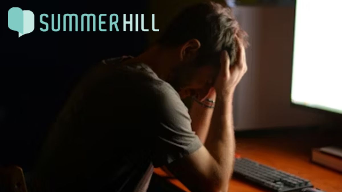 A frustrated man covers his face with his hands, elbows resting on a desk in front of a glowing computer monitor.