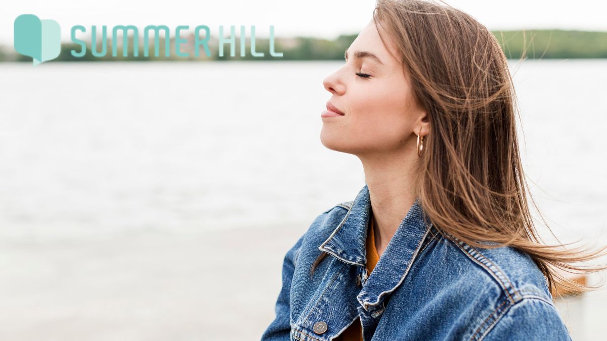 A young woman closes her eyes and takes a deep breath while meditating in front of a scenic ocean view.