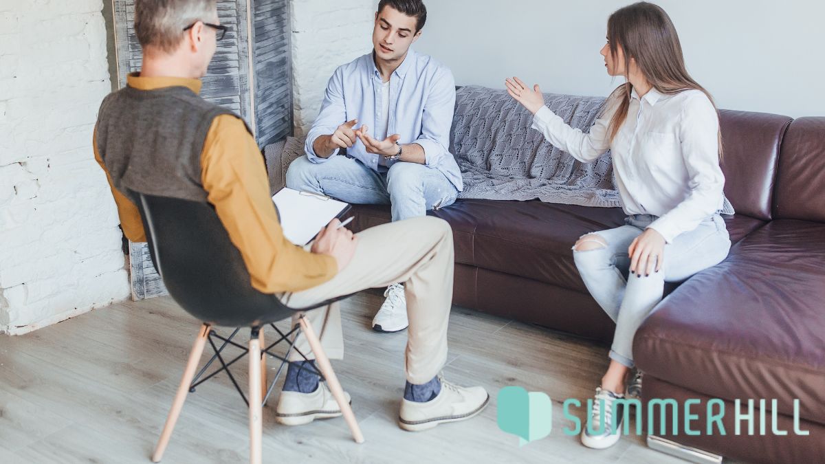 A couple participates in a family counseling session where the woman gestures firmly toward the man, expressing frustration as the therapist observes to guide constructive communication.