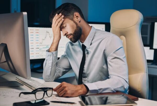 A stressed male executive sits in a dimly lit office, resting his forehead in his hand while facing a computer monitor on a cluttered desk filled with office equipment.