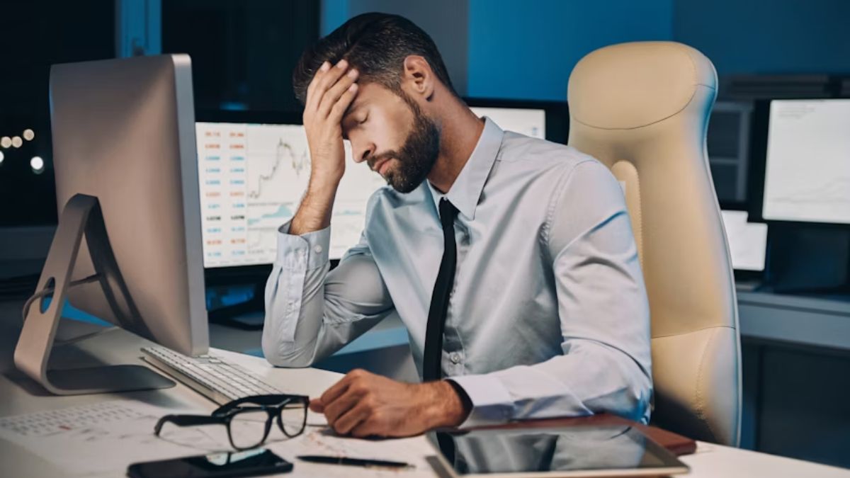 A stressed male executive sits in a dimly lit office, resting his forehead in his hand while facing a computer monitor on a cluttered desk filled with office equipment.